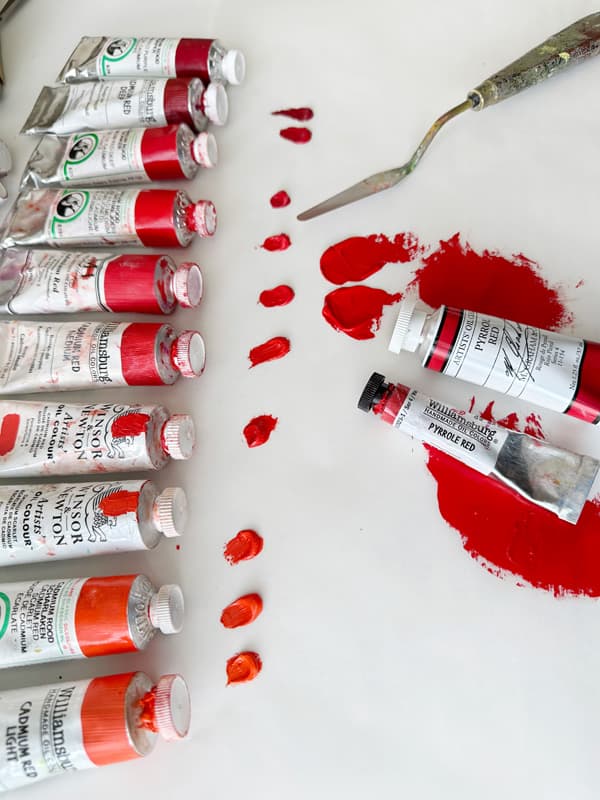 About 10 tubes of cadmium of varying red and red-orange colors with dollops of each paint. Two tubes of Pyrrole Red PR254 are shown. The Pyrrole Reds look most similar to two Cadmium Red Medium colors, one by Michael Harding named just Cadmium Red, and the other close contender was Old Holland's Cadmium Red Vermillioned.
