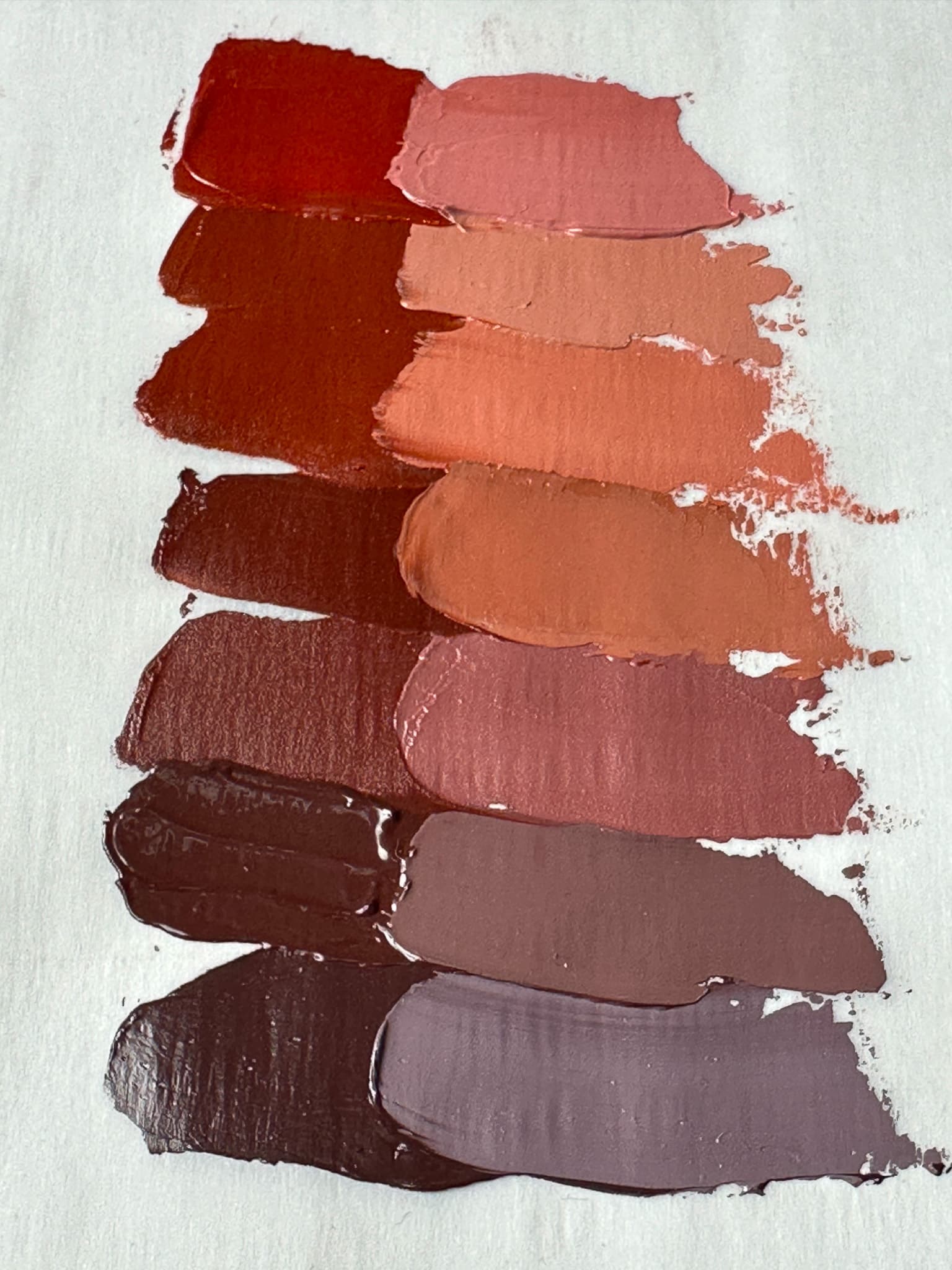 About seven swatches of paint ranging from deep violet brown to maroon to red-orange, which illustrate a range of colors and undertones. All the oil paint swatches are made from versions of PR101